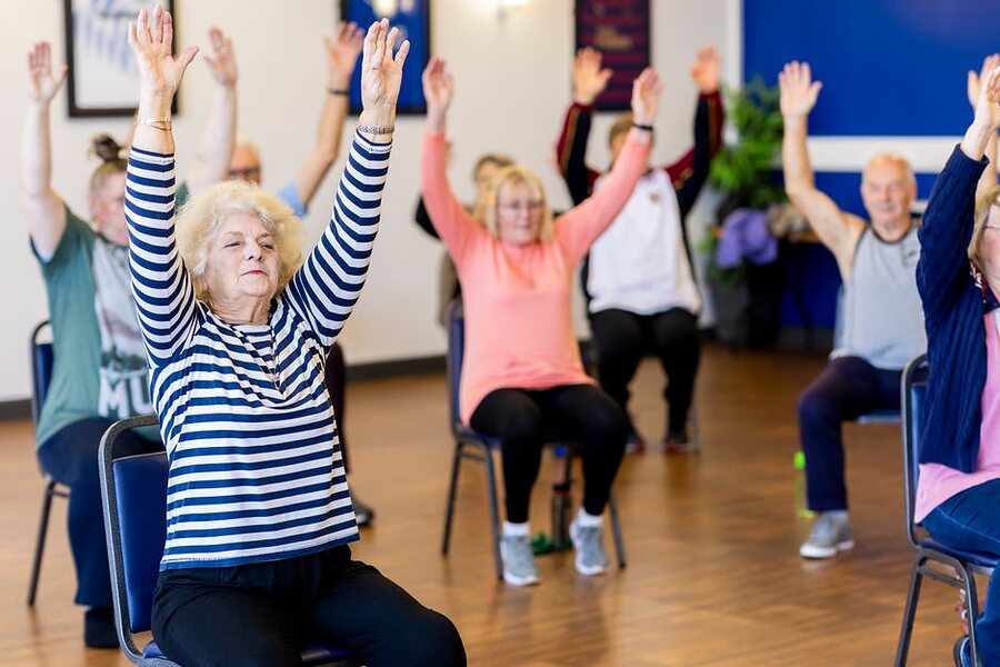 A chair exercise class of older adults, arms raised overhead
